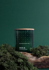 Groene glazen kaarsenpot met een houten deksel, versierd met geometrische patronen. Merknamen "SKANDINAVISK" en "SKOG" label op de voorkant.