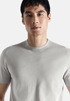 Joven con cabello corto y negro que lleva una camiseta lisa de manga corta y cuello redondo de color gris claro, sobre un fondo blanco.