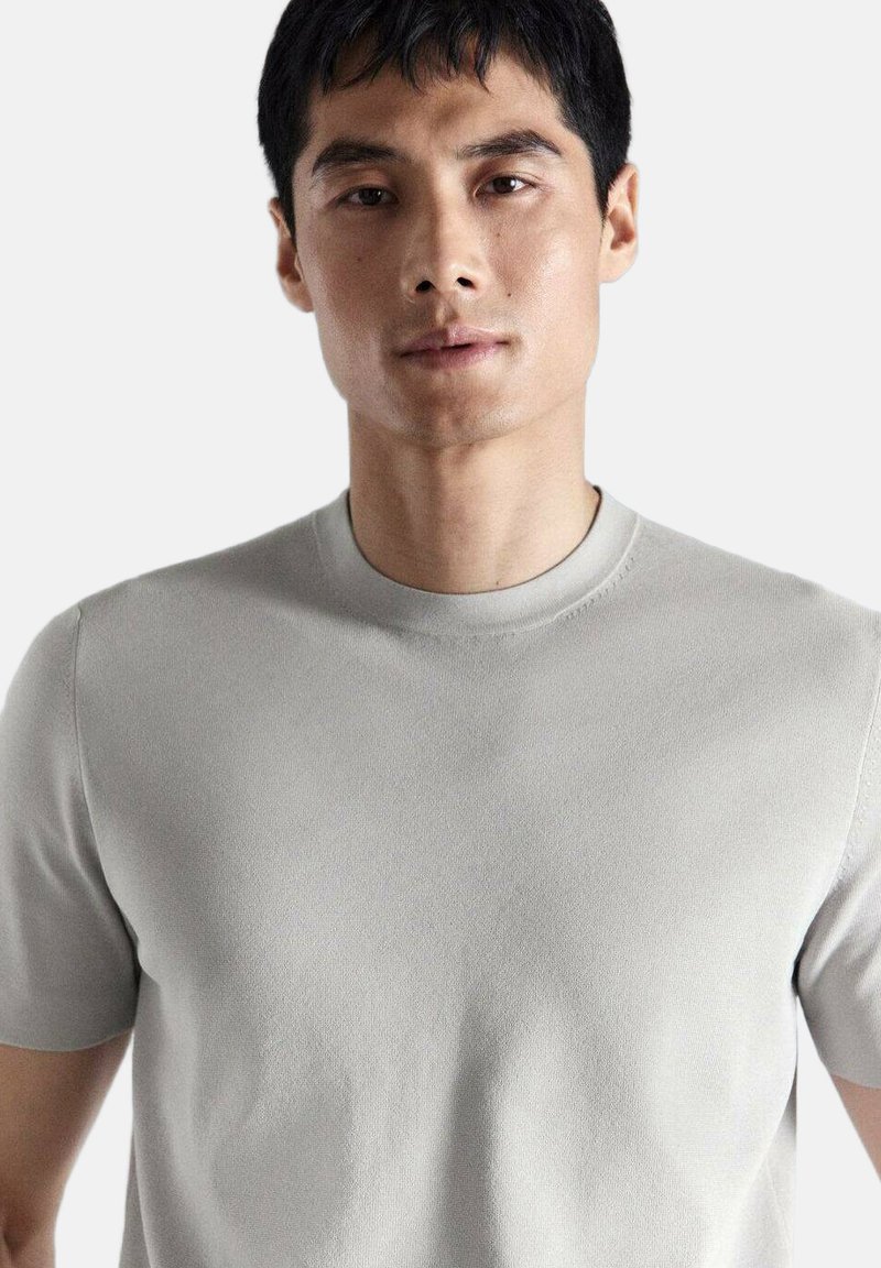 Jeune homme aux cheveux noirs courts portant un t-shirt gris clair uni à manches courtes et col rond, sur fond blanc.