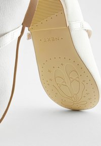 Ballerines blanches en cuir lisse avec une semelle en caoutchouc beige. La semelle présente un design de chaussure de ballet avec des étoiles et un accent en forme de nœud.