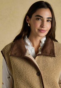 Beige Fleece-Weste mit braunem Cordkragen, Knopfverschluss und strukturierter Oberfläche, über einer weißen Bluse mit Spitzendetails geschichtet.
