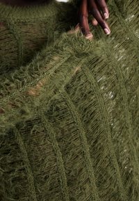 Olivgrüner Strickpullover mit strukturiertem, offenem Muster und fransigen Kanten, der ein weiches, flauschiges Finish und ein geschichtetes Design zeigt.