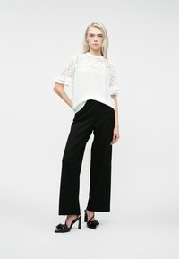 Ξανθιά γυναίκα που στέκεται, φορώντας λευκή δαντελένια μπλούζα με κοντά μανίκια, μαύρους παντελόνες με φαρδιά γραμμή και μαύρες ψηλές γόβες με φιόγκο.