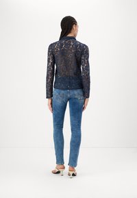 Marineblauw kant lange mouwtop met bloemmotief, semi-doorzichtige achterkant, gecombineerd met lichtblauwe skinny jeans en beige hoge hakken.