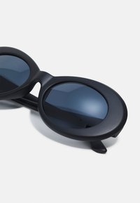 Gafas de sol negras con lentes ovalados y montura mate. Presentan lentes tintados de un azul oscuro, textura suave y patillas delgadas.