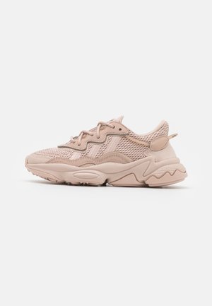 Lyse beige atletisk sneakers med mesh og syntetisk overdel, bølget såldesign og snørebånd set fra siden på hvid baggrund.