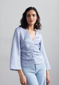 Lyseblå, teksturert bluse med V-hals og rynket front. Har lange klokkeermer og er parret med lyseblå jeans.