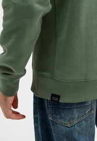 Groene sweatshirt met ribgebreide manchetten en onderrand, gecombineerd met blauwe jeans met een versleten textuur en een zaklabel met de tekst "Butcher of Blue."