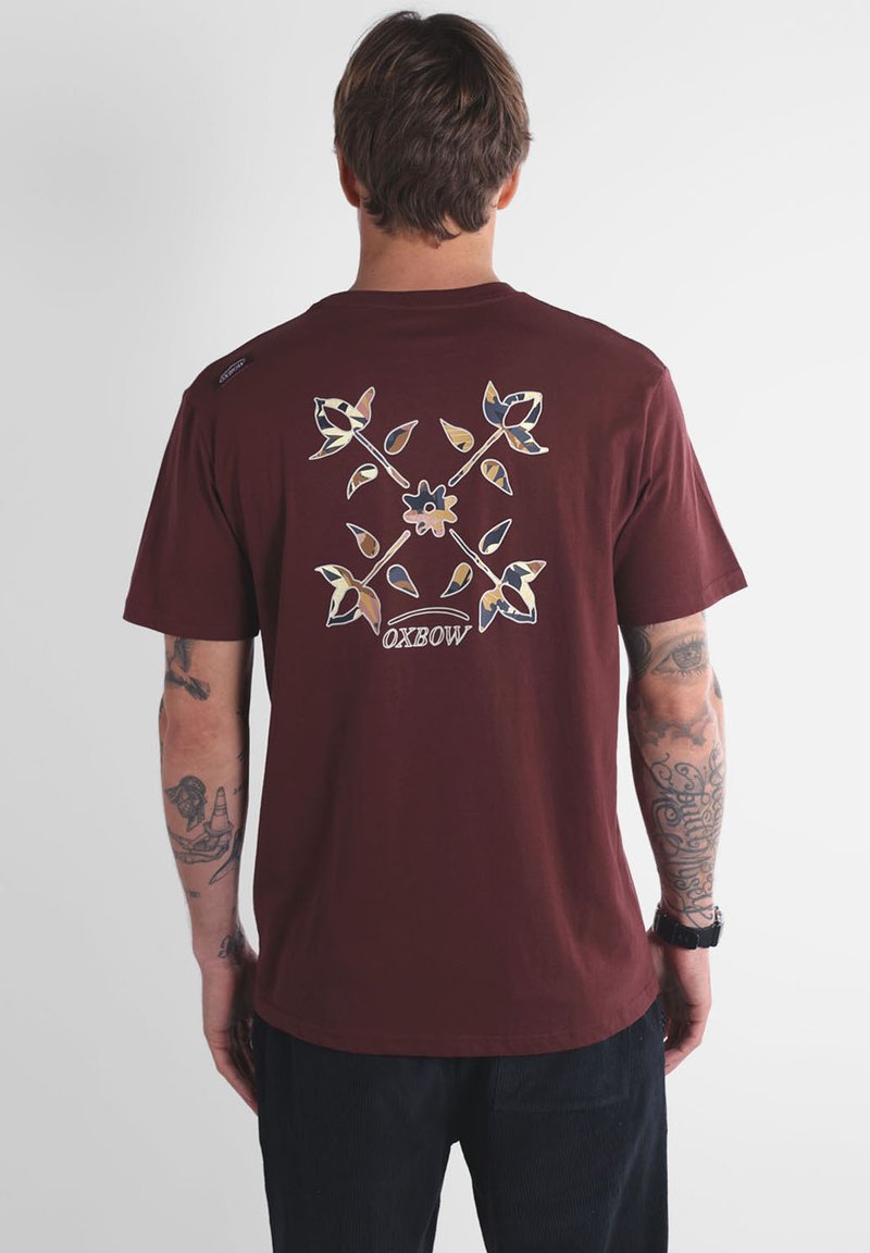 Homme avec des tatouages portant un t-shirt bordeaux orné d'un design floral et de feuilles avec le texte "OXBOW" à l'arrière, se tenant devant un fond uni.