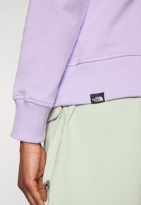 Felpa viola chiaro con polsini a costine e etichetta nera di The North Face, indossata sopra un indumento verde. Tessuto morbido e texturizzato.