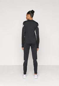 Čierne športové tričko s dlhým rukávom a priliehavé nohavice, s bielymi diagonálnymi pruhmi na ramenách a spodnej časti nohavíc. Obuv tvoria svetlé tenisky.