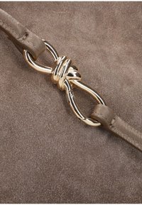 Leren band in tan kleur met een gouden metalen knoopdetail. De achtergrond heeft een gestructureerd taupe oppervlak, dat glad aanvoelt tegen de afwerking van de band.