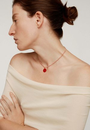 Mujer con el cabello recogido en un moño, vistiendo una blusa beige con hombros al descubierto y un collar de cuentas rosas con un colgante en forma de estrella roja.