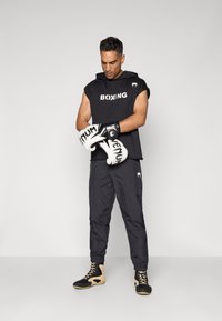Sort ærmeløs hoodie med "BOXING" print, parret med sorte træningsbukser. Hvide bokseshandsker med "VENUM" logo. Sorte og tan atletiske sko.
