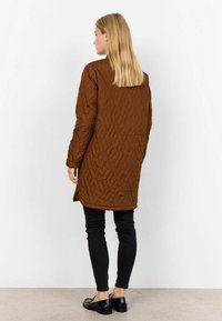 Brun kviltad lång kappa med en texturerad yta, tillsammans med svarta åtsittande byxor och svarta loafers, sedd från baksidan.