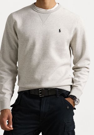 Uomo che indossa una felpa girocollo grigio chiaro con piccolo logo polo nero, pantaloni cargo neri con cintura e orologio da polso, una mano in tasca.