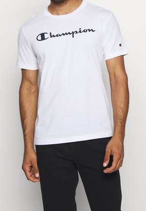 Muž oblečený v bílém tričku značky Champion s černým logem a černými kalhotami, stojící uvolněně na pozadí s jednobarevným podkladem.