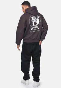 Dunkelgrauer, übergroßer Hoodie mit Kapuze und grafischem Druck auf der Rückseite. Kombiniert mit schwarzen, lässig sitzenden Hosen und weißen Sneakern.