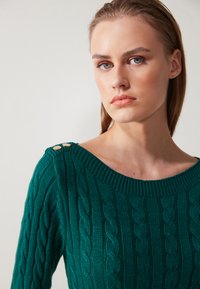 Green knitted sweater featuring a cable pattern, wide neckline, and button accents on the shoulder. Smooth texture with ribbed cuffs.