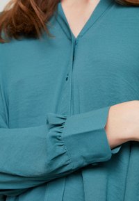 Blusa verde acqua con una consistenza morbida, caratterizzata da un collo a V, bottoni e polsini arricciati. Il tessuto cade delicatamente e ha una finitura liscia.