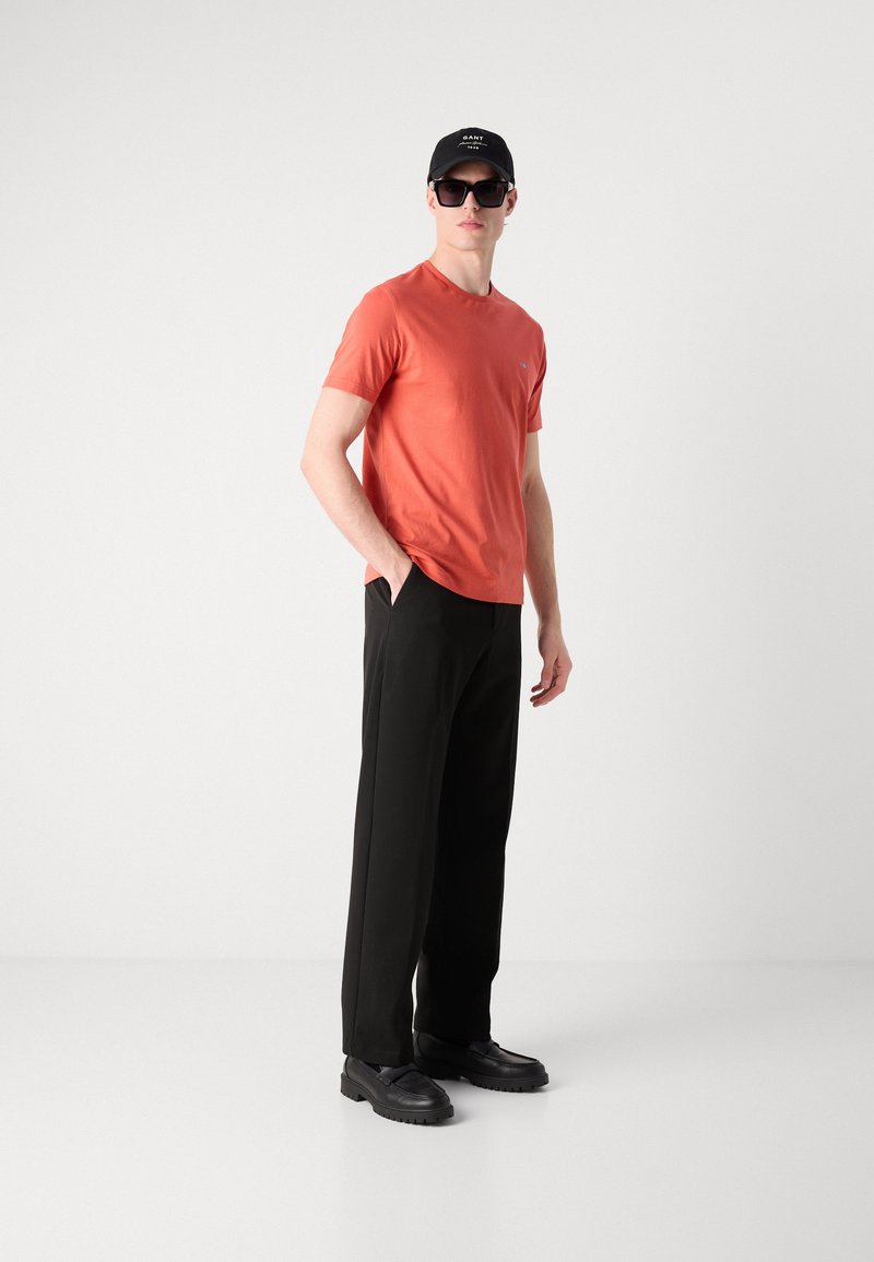 Orange T-Shirt mit kurzen Ärmeln und rundem Ausschnitt, schwarze weite Hose, schwarze klobige Schuhe und schwarze Mütze mit weißem Logo.