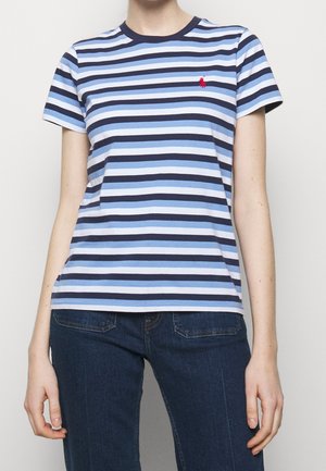 Woman wearing a short-sleeve blue and white striped t-shirt with a small red logo on the chest and dark blue jeans.