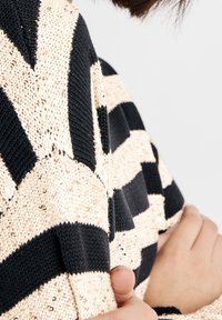 Gebreide trui met een zigzagpatroon in zwart en beige, met een gestructureerde, sprankelende afwerking en een losse pasvorm rond de schouders.