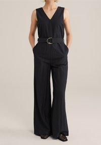 Czarny bezrękawnik z dekoltem w szpic, talią ściągniętą paskiem z okrągłą złotą klamrą oraz szerokimi spodniami. Tkanina o fakturze.