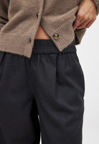 Bruine wollen cardigan met geribbelde zoom en twee knopen, gecombineerd met zwarte, geplooide broek met elastische taille. Textuurdetails van de stof zichtbaar.