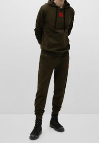 Olivově zelená mikina s kapucí s červenou nášivkou loga, doplněná o odpovídající tepláky; oba mají měkkou texturu, boční kapsy a žebrované manžety.