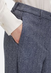Marineblauwe herringbone broeken met een gestructureerd patroon, met een aansluitende taille en zijzakken. Close-up van een hand die in de zak rust.