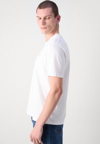 Witte katoenen t-shirt met korte mouwen, ronde hals en een subtiel logo op de borst. Draagt bij blauwe denim jeans.