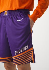 Fioletowe szorty do koszykówki z pomarańczowymi akcentami, z siateczkową teksturą, elastycznym paskiem w talii i bocznymi rozcięciami. Na nich w białym kolorze napis "PHOENIX".