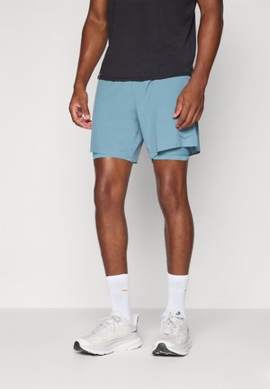 Shorts de sport bleu clair à coupe ample avec poches latérales, associés à un t-shirt noir, des chaussettes blanches et des baskets de couleur claire.
