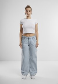 Mladi žena s kosom u punđi, obučena u bijelu kratku majicu, svijetloplave široke traperice, bijele tenisice i zlatne naušnice, stoji uspravno.