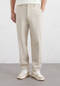 Beige Baumwollhose mit geradem Schnitt, ausgestattet mit Fronttaschen und einer glatten Textur, kombiniert mit weißen Sneakern.