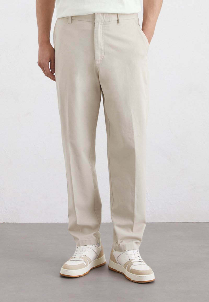 Beige Baumwollhose mit geradem Schnitt, ausgestattet mit Fronttaschen und einer glatten Textur, kombiniert mit weißen Sneakern.