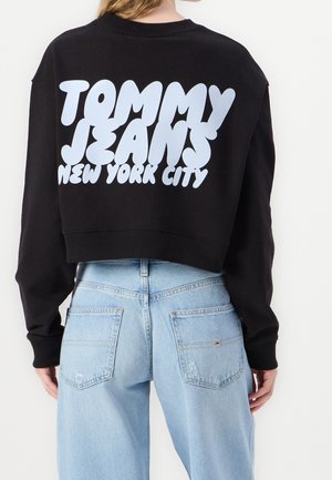 Osoba ubrana w czarną krótką bluzę z napisem „Tommy Jeans New York City” i jasnoniebieskie jeansy z wysokim stanem, widziana od tyłu.