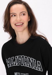Fekete pulóver, melyen fehér, vastag betűkkel olvasható a "CALIFORNIA" felirat, alatta pedig a "WEST COAST" felirat található; kerek nyakú dizájn, puha anyag, laza fazon.