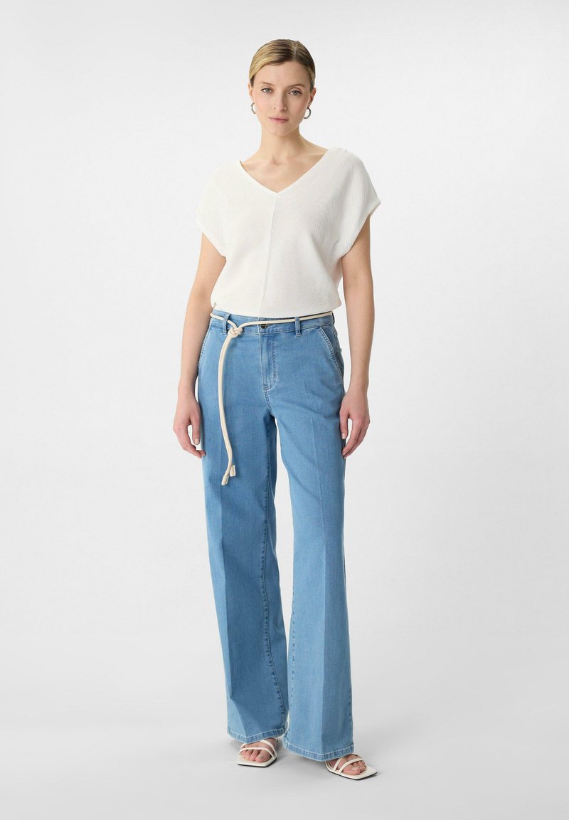White ribbed short-sleeve top with a V-neck, paired with light blue high-waisted flared jeans and a beige belt. Model wears white sandals.