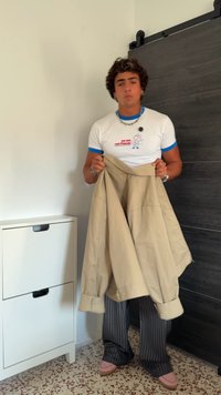 Casaco bege segurado nas mãos, com grandes bolsos e punhos dobrados. O modelo usa uma t-shirt branca com detalhes azuis e calças cinza risca de giz.