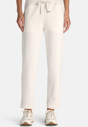 Kvinde iført off-white bukser med snøre i taljen og frontlommer samt matchende off-white sneakers, stående mod en ensfarvet baggrund.