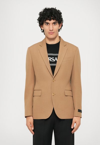 Blazer marron sur mesure avec un design structuré, deux poches avant et des boutons dorés. Porté sur un t-shirt graphique noir. Texture de tissu lisse.