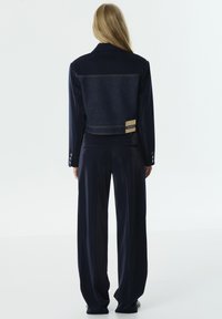 Marineblauwe cropped jacket met een denim achterkant, een getailleerde structuur en lange, rechte broekspijpen met een gepolijste textuur. Label aan de achterkant.