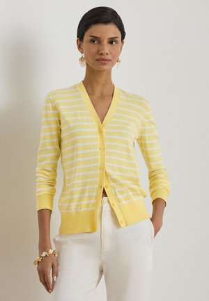 Žena v žluto-bílém pruhovaném kardiganu s výstřihem do V, bílých kalhotách, náušnicích ve tvaru lastury a náramku ladícím s náušnicemi, stojící u jednobarevné stěny.