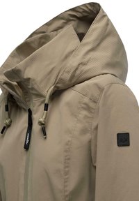 Gai bēšas krāsas jaka ar kapuci, kas izceļas ar ribotu tekstūru, sažņaugšanas detaļām un melnu logo uzlikumu uz piedurknes. Ar rāvējslēdzēju aizdare priekšpusē.