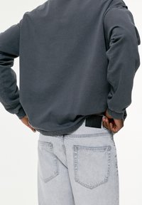 Felpa grigia con vestibilità comoda, abbinata a jeans di denim grigio chiaro. Caratteristiche includono polsini a costine e un'etichetta parzialmente visibile nella vita.