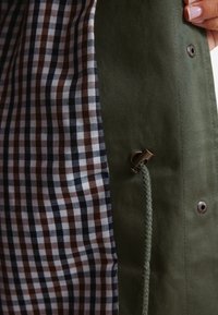 Olivengrøn stoffer coat med en brun og navy rutet foring. Har en metal trykknap og en flettet snor detalje.