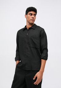 Svart skjorte med smale striper, lange ermer, knappestolpe foran og brystlomme; laget av et glatt, lett materiale.