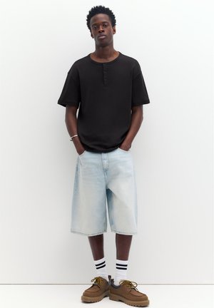 Jeune homme debout contre un mur blanc, portant un maillot henley noir à manches courtes, un short long en denim clair, des chaussettes blanches à rayures noires et des chaussures marron.